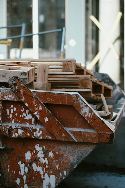 A close-up view of a rusted metal boat filled with several wooden pallets arranged in neat stacks, positioned on a paved surface near a building with large glass windows and metal framework. The woode