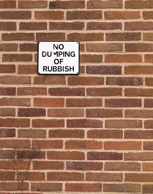 A white rectangular sign with black text reading 'NO DUMPING OF RUBBISH' is mounted on a red brick wall. The wall consists of uniformly arranged bricks with a rough texture, in shades of reddish-brown